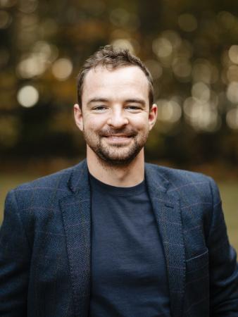 Patrick Tolosky smiling and standing outside in front of a wooded backdrop