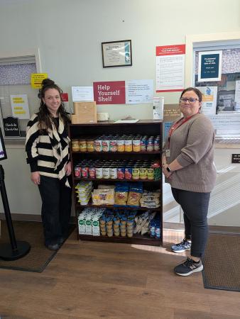 Two women standing next to a shelf stocked with non-perishable food items 