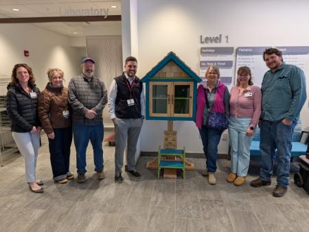 A group of adults standing together smiling next to a small house-shaped box containing books. They are celebrating together.