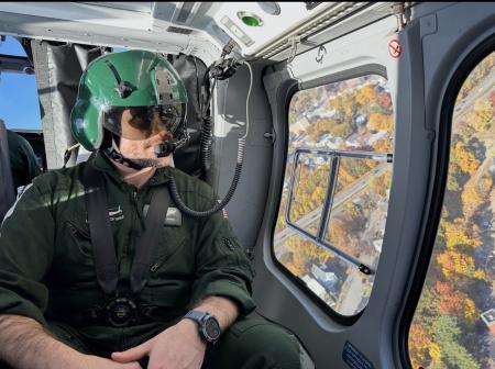 close-up of a man wearing a flight suit and helmet in a helicopter as it flies over a rural community below