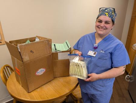 A woman in scrubs smiling and holding bags of donated breast milk standing next to at table with a large brown box on it