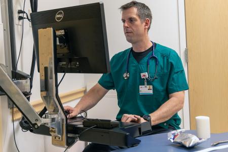 A man wearing teal scrubs, standing at a computer terminal in a medical office