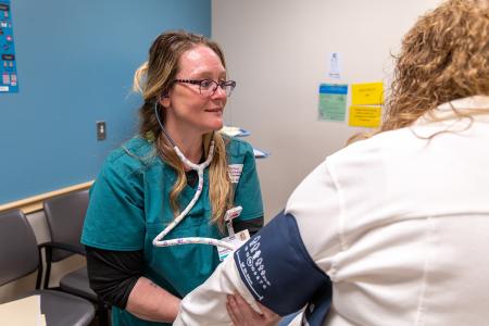 A woman wearing teal scrubs is sitting in a medical exam room. She is using a stethoscope and blood pressure cuff to take a patient's blood pressure reading