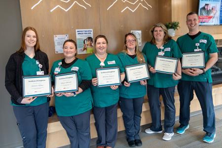 A group of adults wearing matching teal scrubs, standing together in a hallway holding up diplomas and smiling