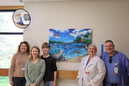 A group of five adults standing in front of a painting hanging on a hospital wall. The painting is of a serene ocean scene with lots of blue water and skies.