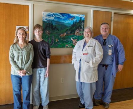 Four adults standing together and smiling next to a painting hanging on a hospital wall. The painting is of a peaceful woodland scene with bright green grass and blue skies