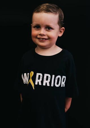 A smiling boy stands in front of a black background wearing a shirt that reads "Warrior"