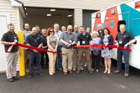 a group of adults participating in a ribbon cutting ceremony outside a building and next to an ambulance