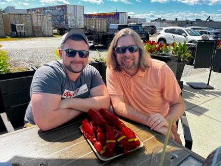 Two men sitting together outside and smiling with a tray of cooked lobsters in front of them