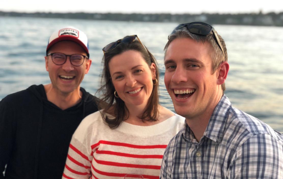 Three adults (Susan, Grant, and Brett) standing together and smiling in front of a large body of water.