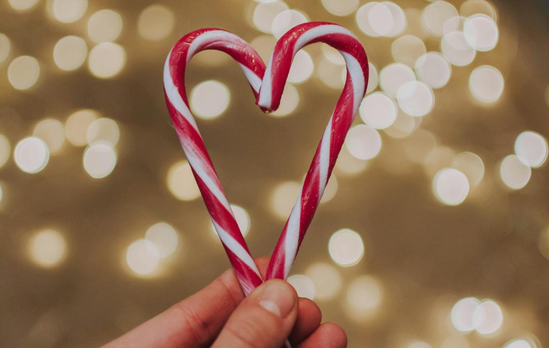A hand holding two candy canes forming a heart shape with soft sparkle, twinkle lights behind them