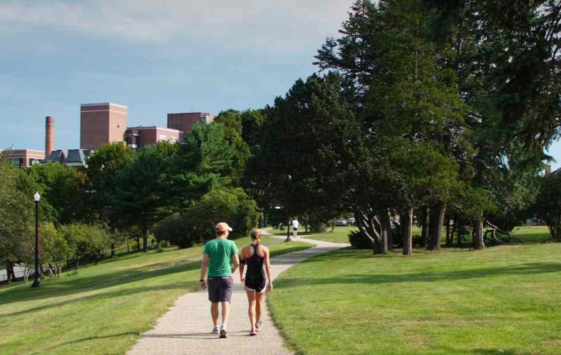 Two people in workout clothes walk together on a path surrounded by grass and trees
