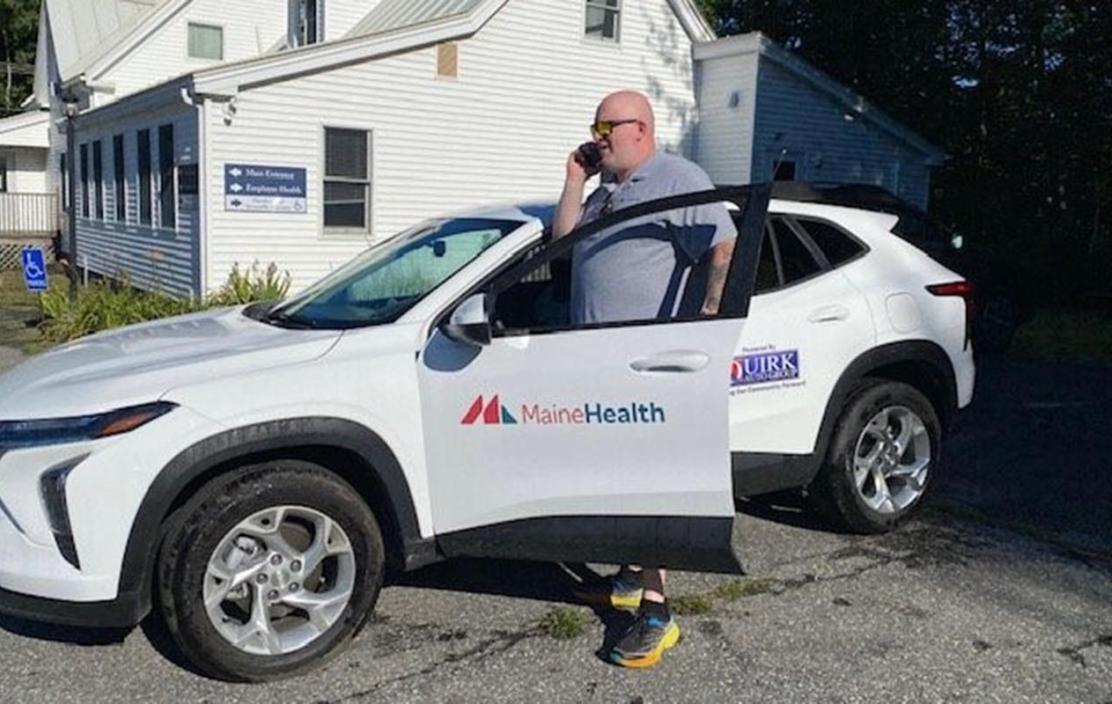 A man is standing outside next to a white car. The door has a MaineHealth logo on it and is open slightly. The man is a community health work who is on the phone and speaking with a client he is to visit.