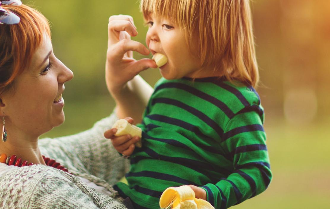 A women feeds her child a piece of banana outside.