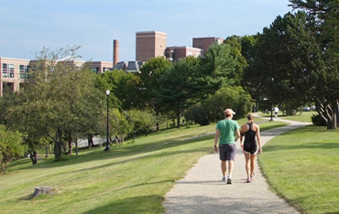 Two people walk on a path surrounded by green grass and trees. Maine Medical Center is seen in the distance.