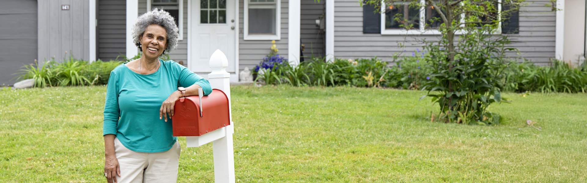 An older woman in a teal shirt, smiling as she stands next to her bright red mailbox and rests her elbow on it.