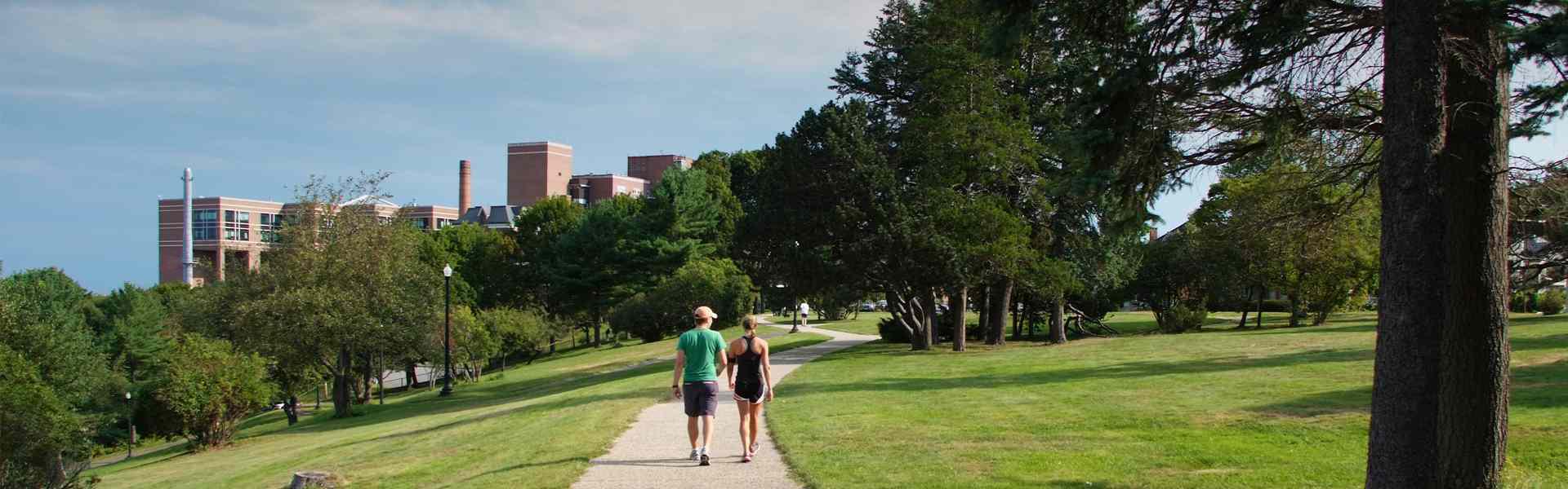 Two people in workout clothes walk together on a path surrounded by grass and trees