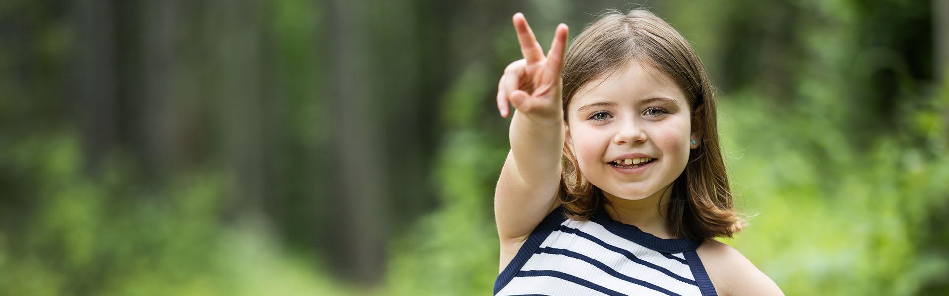 A young girl standing in the woods and smiling. She is holding her hand up in a "peace" sign.
