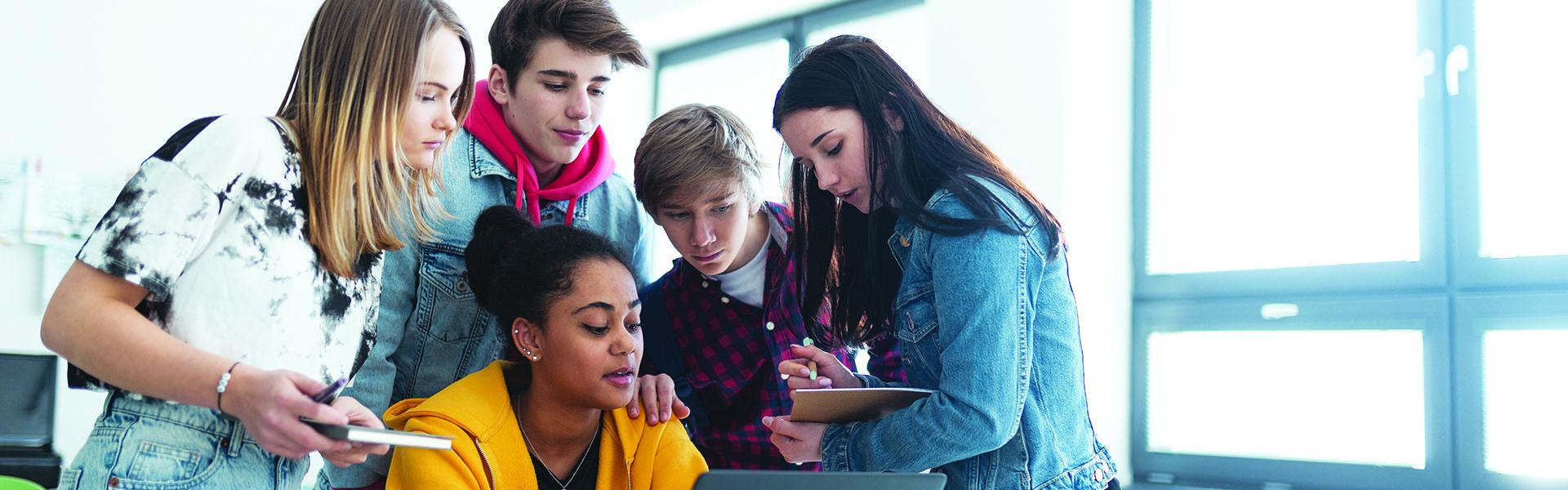 a group of teenage student sitting together looking at a laptop screen in a classroom setting