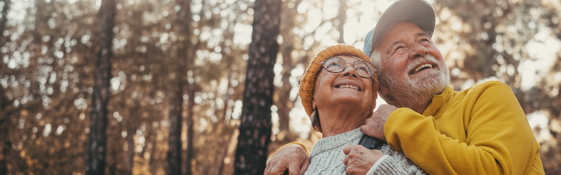 Two older adults outside in a forest on a sunny day. The man is resting his hand on the woman's shoulders affectionately as both are looking upwards with a smile