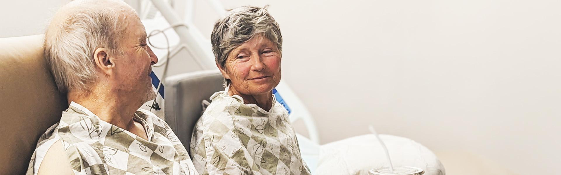 An elderly couple sits side by side in hospital beds, both wearing patterned hospital gowns. The man on the left smiles warmly as he looks toward the woman, while she smiles back at him with a gentle expression.