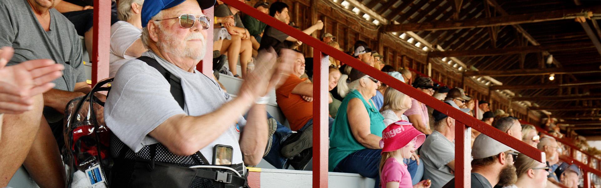An older man wearing a blue cap, sunglasses, and an LVAD device harness claps from the wooden grandstands at a community event, surrounded by a crowd of all ages.