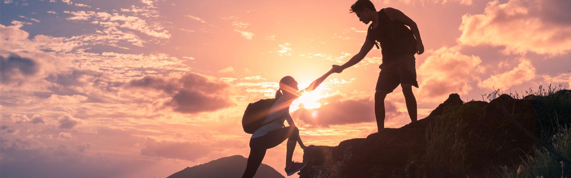 Two hikers helping each other up a mountain are silhouetted against a sunset