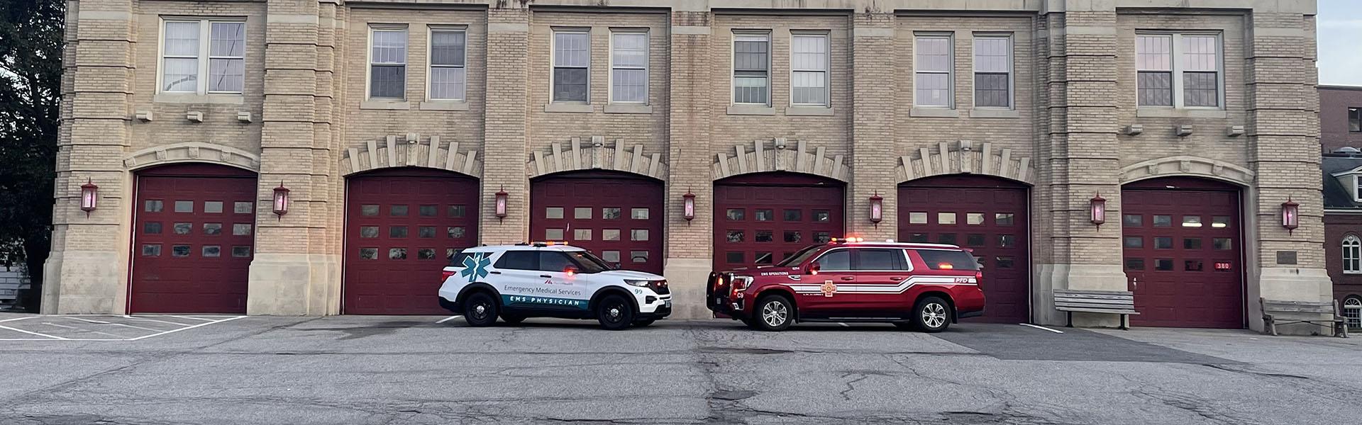 Two emergency medical services SUVs parked nose-to-nose in front of a line of red fire station doors