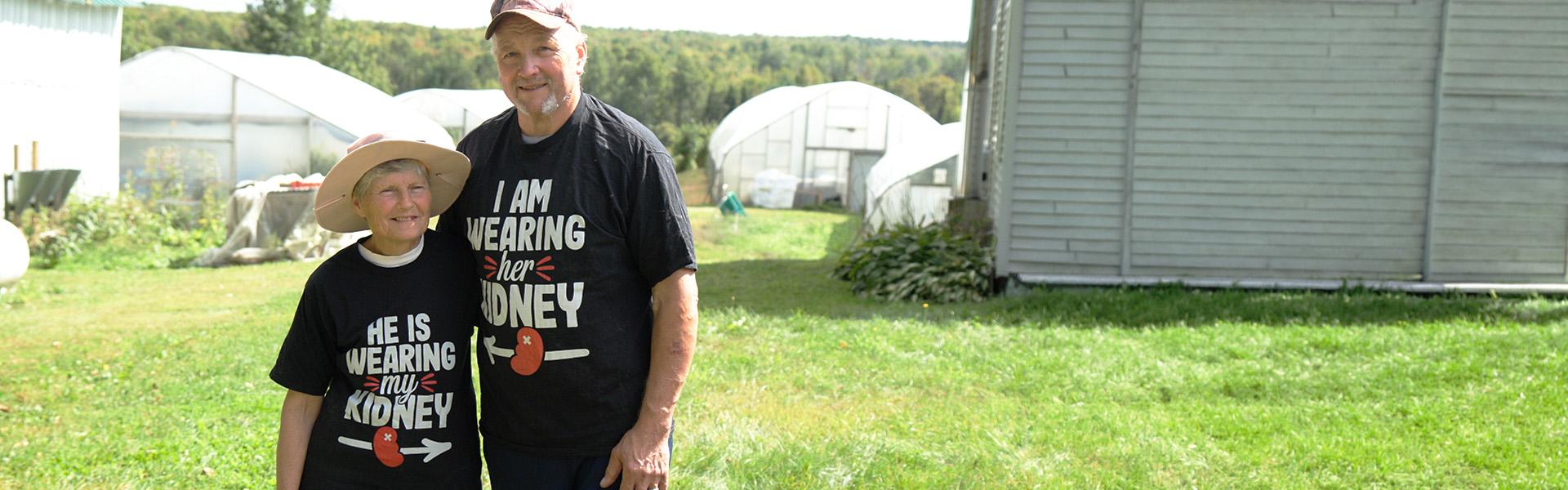 A kidney donor and recipient couple stand smiling together on a farm, each wearing matching black t-shirts — hers reading "He is wearing my kidney" and his reading "I am wearing her kidney.