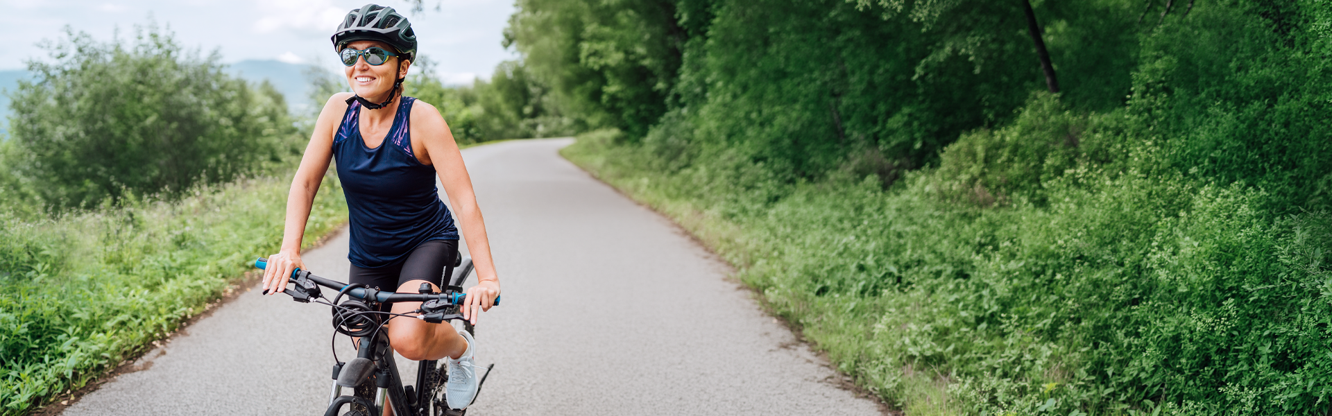 A woman smiling as she rides her bicycle on a paved trail through the woods