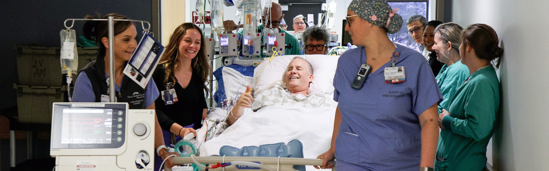 MaineHealth care team members move a smiling patient on a gurney who is giving the "thumbs up" sign