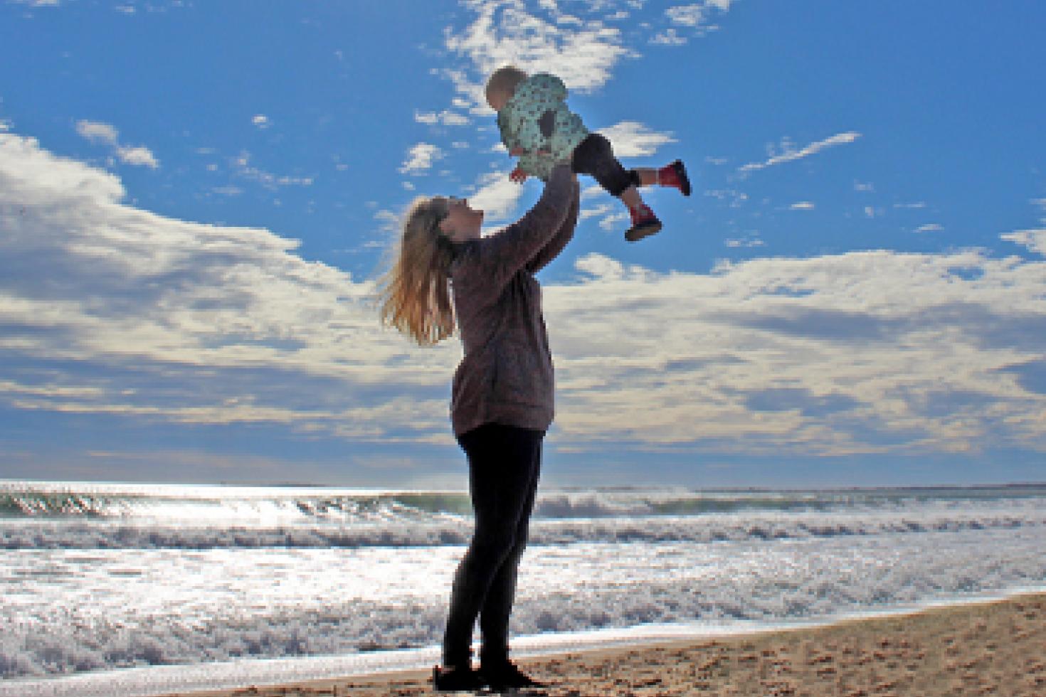 A mother stands on a beach holding a child in the air above her head