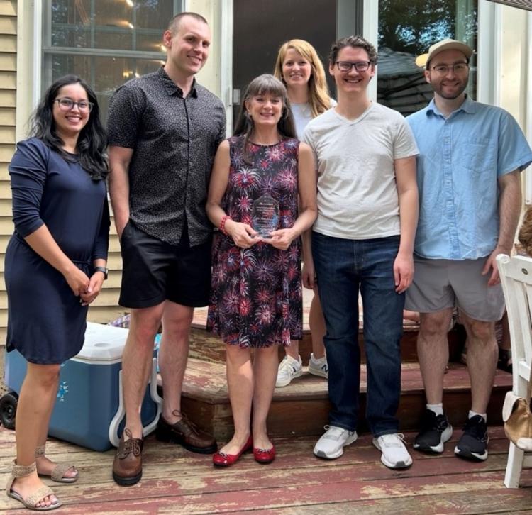 Five hematology fellows stand together on a brick patio. One faculty member stands in the center of the group, holding an award.