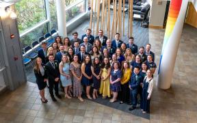 A group of the Maine Track graduates standing together in a bright, sunny atrium on their graduation day