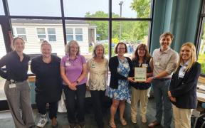A group of adults standing together and smiling in front of a large window. One is proudly holding a certificate.