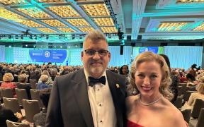 Devin Carr and Valerie Fuller dressed formally and standing together at an awards ceremony in a large room full of people