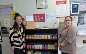 Two women standing next to a shelf stocked with non-perishable food items 