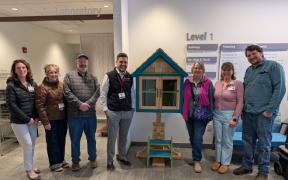A group of adults standing together smiling next to a small house-shaped box containing books. They are celebrating together.