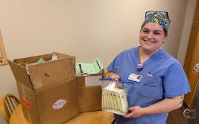 A woman in scrubs smiling and holding bags of donated breast milk standing next to at table with a large brown box on it