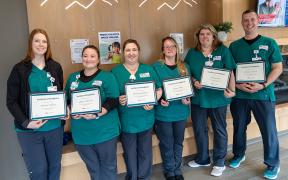 A group of adults wearing matching teal scrubs, standing together in a hallway holding up diplomas and smiling