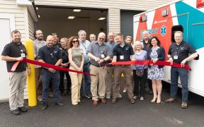 a group of adults participating in a ribbon cutting ceremony outside a building and next to an ambulance