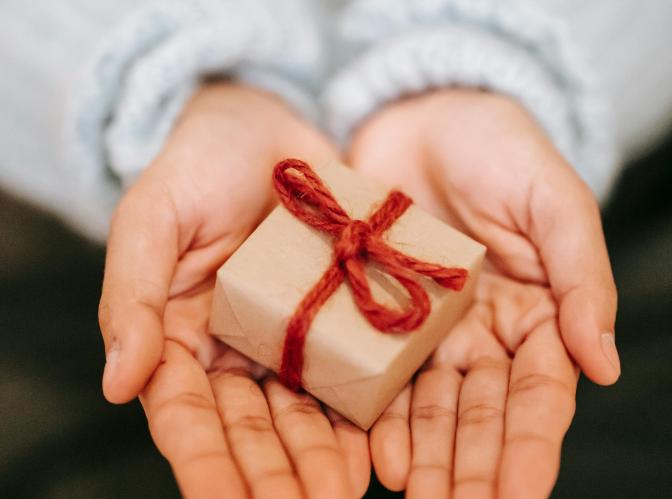 A pair of hands extended with their palms up, in them is a small gift box wrapped in light brown paper and tied with a red string
