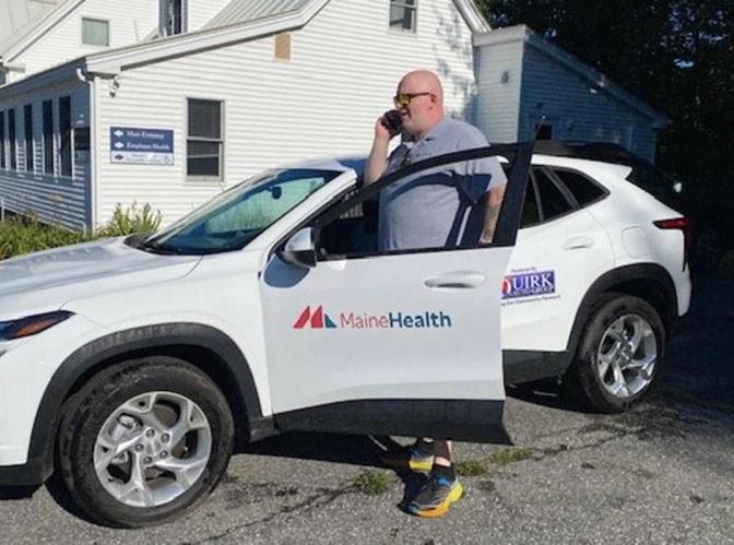 A man is standing outside next to a white car. The door has a MaineHealth logo on it and is open slightly. The man is a community health work who is on the phone and speaking with a client he is to visit.