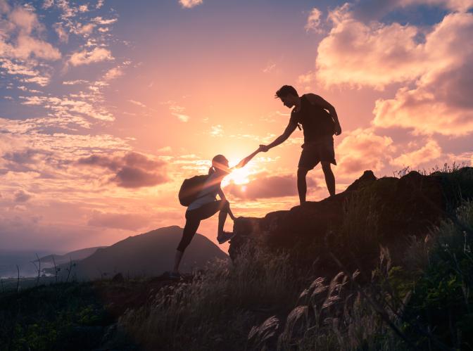 Two hikers helping each other up a mountain are silhouetted against a sunset