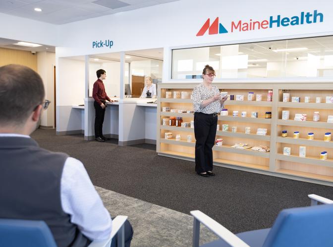 Interior of the MaineHealth Pharmacy on Brighton Ave in Portland - a man is being helped at the counter, while a woman peruses various health care items on shelves nearby