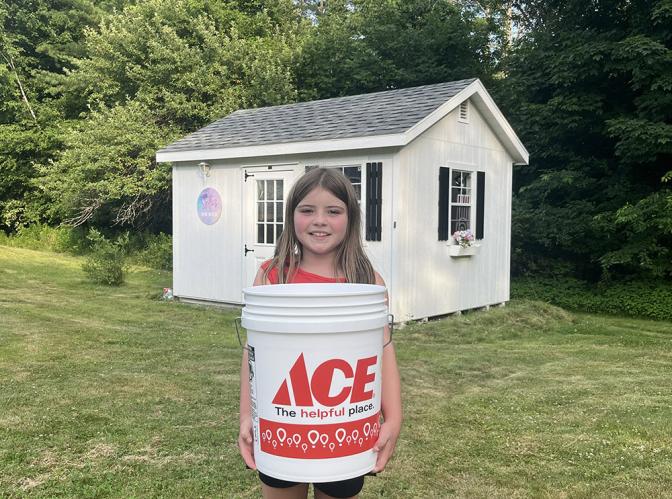 A young girl with a big smile, holding a large "ACE" branded construction bucket. She is standing outside in her backyard on a bright day.
