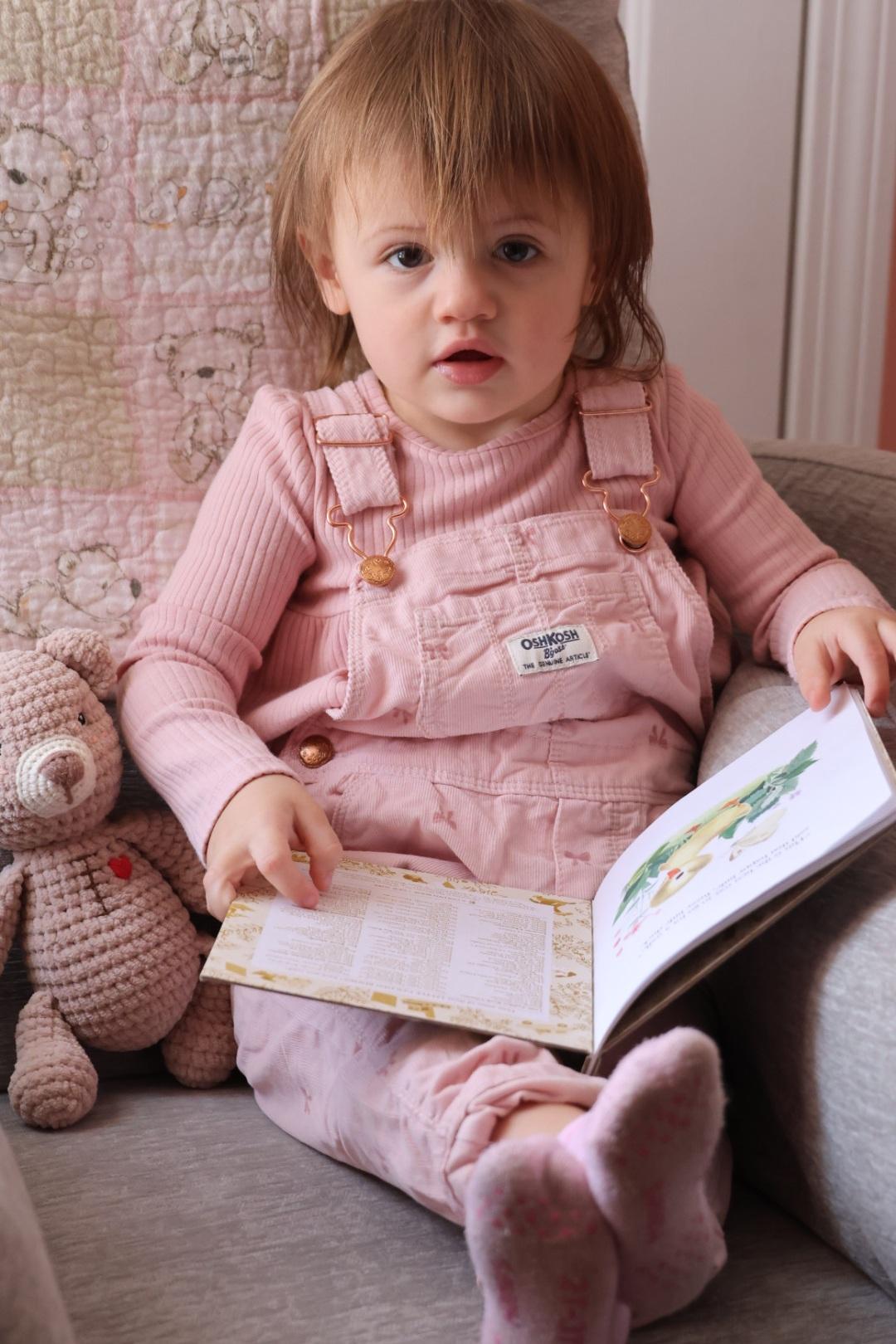 A young girl wearing pink overalls, reads a book next to her teddy bear