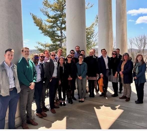 A group of adults standing together outside next to large white columns, a blue sky and trees are behind them