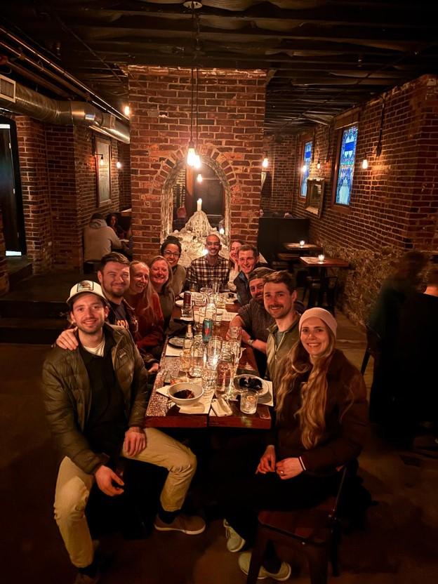 a group of adults sitting together around a large dining table in a brick-walled restaurant. They are smiling and having a good time together.
