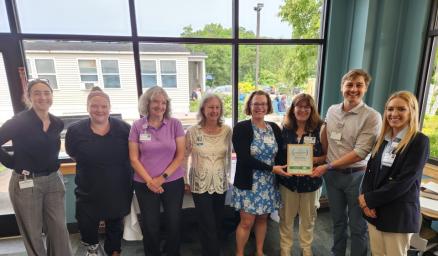A group of adults standing together and smiling in front of a large window. One is proudly holding a certificate.